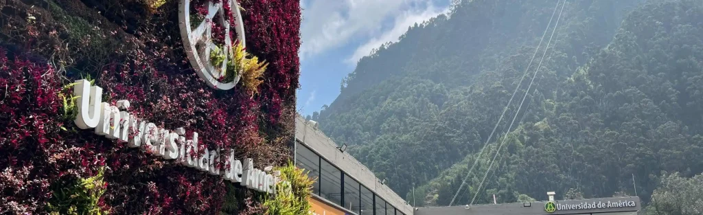 Fachada de la Universidad de América en Bogotá con muro verde y montañas al fondo.